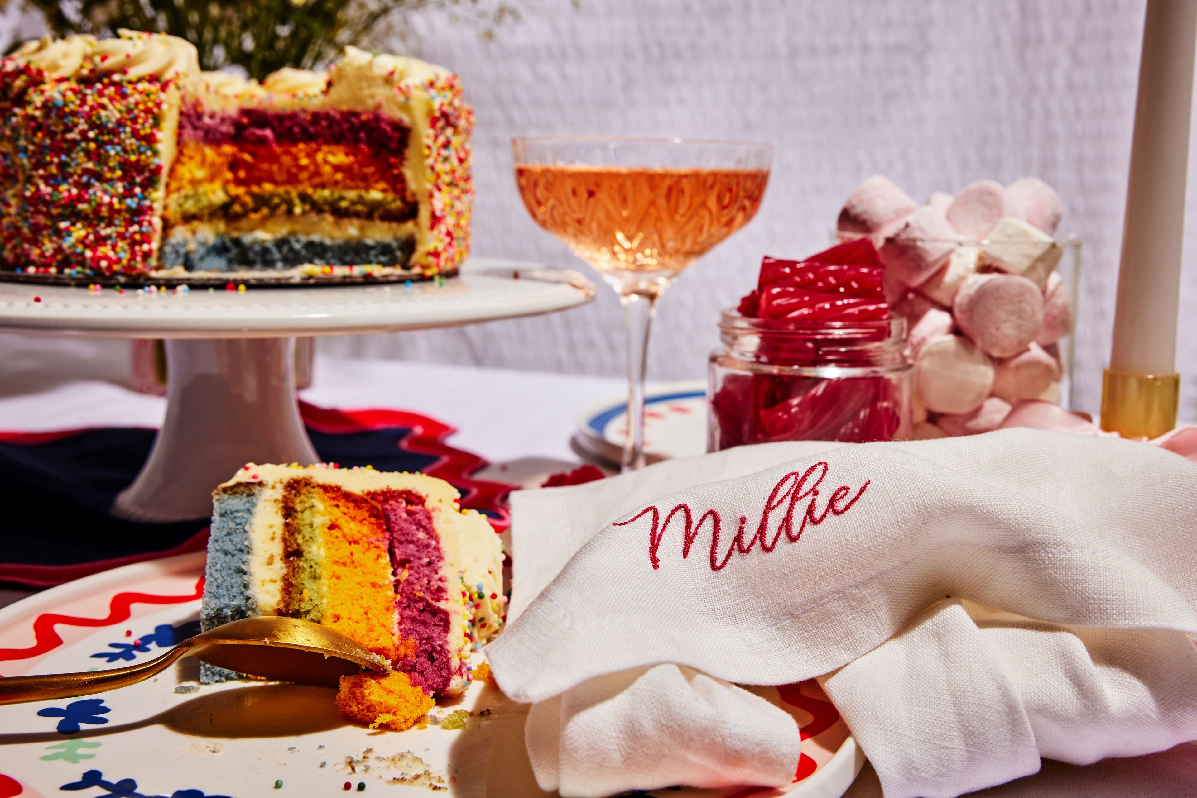 Custom white linen napkin with “Millie” embroidery, styled beside rainbow cake, pink sweets, and sparkling rosé