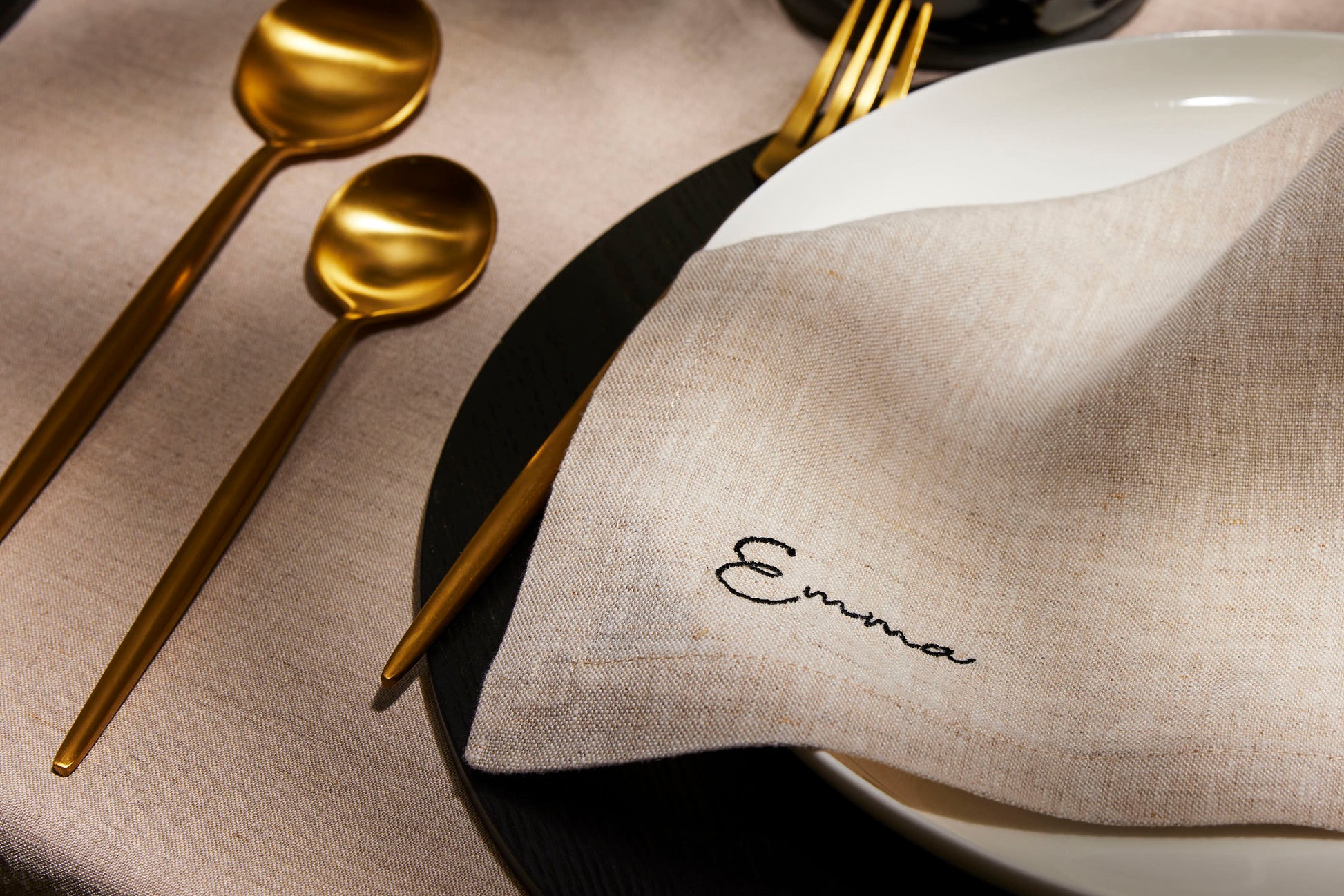 Beige linen napkin embroidered Emma in black, styled with gold cutlery and dark-toned tableware